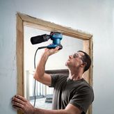 Một người mặc đồ bảo hộ đang khoan vào khung cửa gỗ bằng một dụng cụ điện.