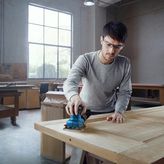 Một người mặc đồ bảo hộ đang chà nhám cửa gỗ bằng máy chà nhám cầm tay.