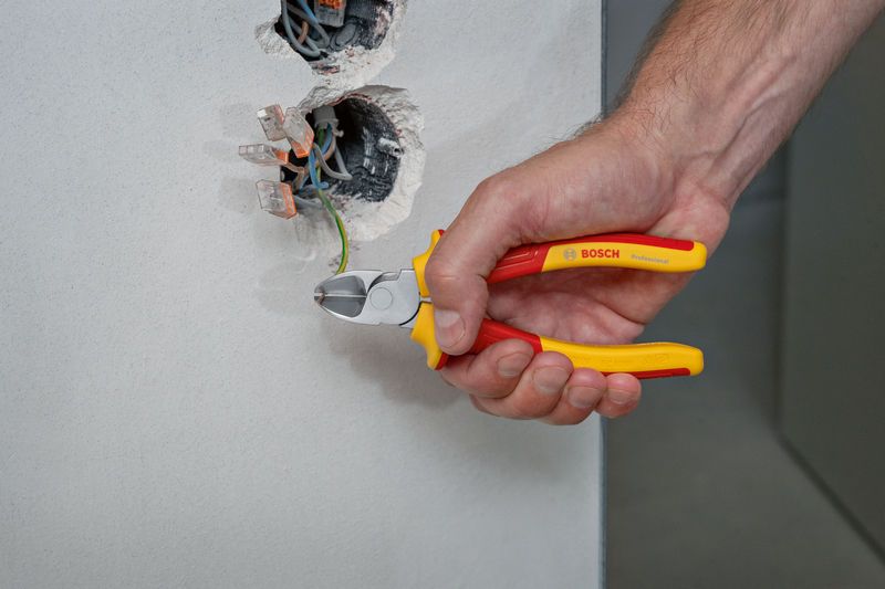 Kìm cắt chéo bên VDE 160mm Một người sử dụng kìm cách điện để cắt dây màu xanh lá cây từ hộp điện trên tường.