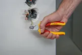 Một người sử dụng kìm cách điện để cắt dây màu xanh lá cây từ hộp điện trên tường.