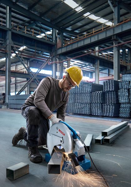 Một người mặc đồ bảo hộ đang cắt ống kim loại bằng máy cưa trong một nhà kho.