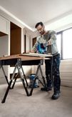 Một người mặc đồ bảo hộ đang sử dụng máy cưa lọng để cắt gỗ trên bàn làm việc.