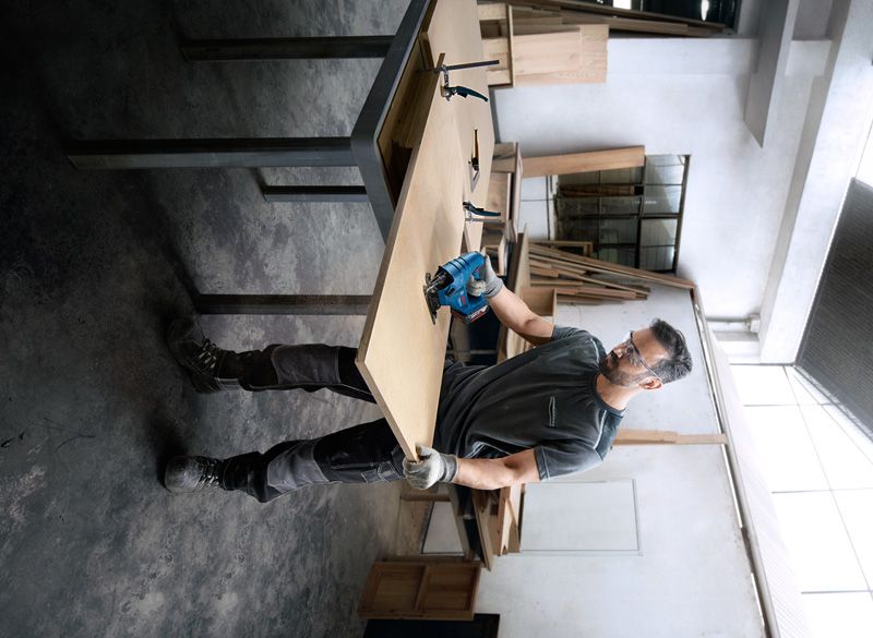 GST 183-LI Một người mặc đồ bảo hộ đang cắt gỗ bằng dụng cụ điện trong xưởng.