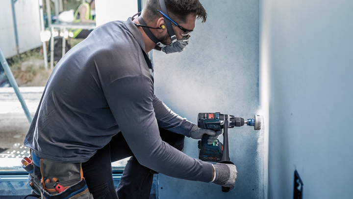 Một người mặc đồ bảo hộ đang khoan một lỗ tròn trên tấm vách thạch cao.