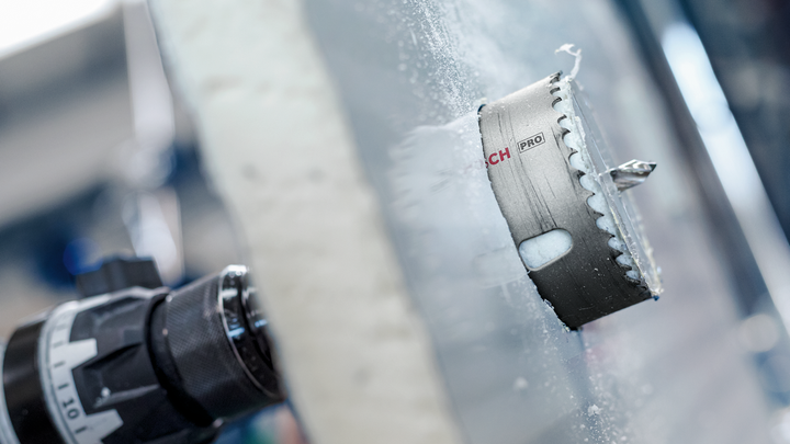 Lưỡi cưa lỗ gắn vào máy khoan đang cắt qua bề mặt kính mờ.