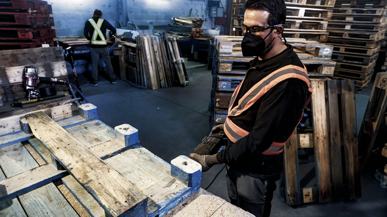 Một người mặc đồ bảo hộ đang sử dụng dụng cụ điện để làm việc trên một tấm pallet gỗ.