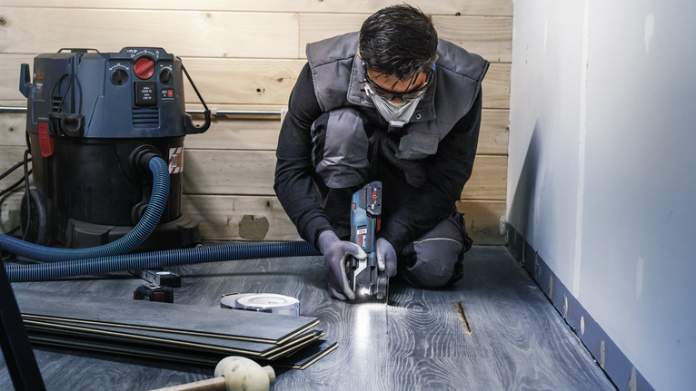Một người mặc đồ bảo hộ đang cắt sàn gỗ công nghiệp bằng dụng cụ điện.