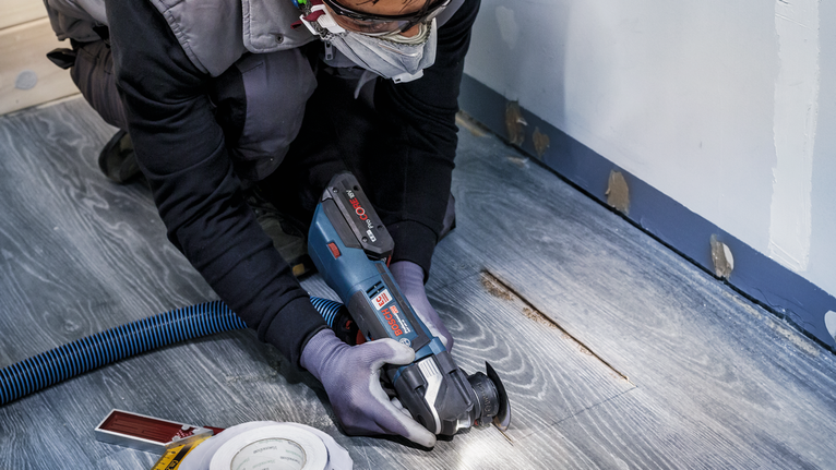 Người mặc đồ bảo hộ đang cắt sàn gỗ bằng dụng cụ điện.