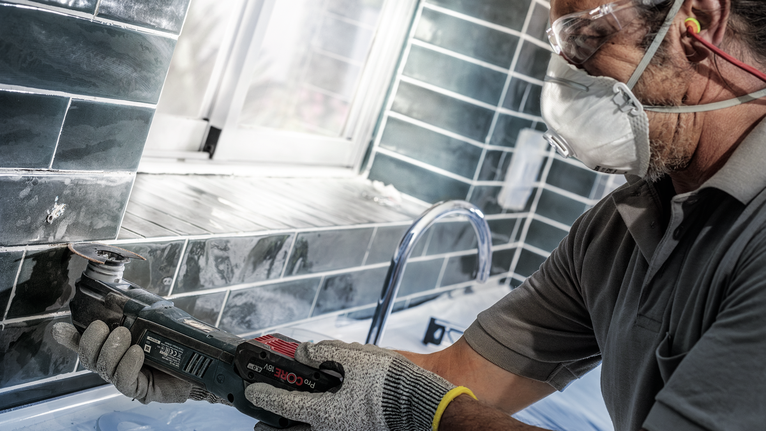 Một người mặc đồ bảo hộ đang sử dụng dụng cụ điện để cắt gạch gần bồn rửa.