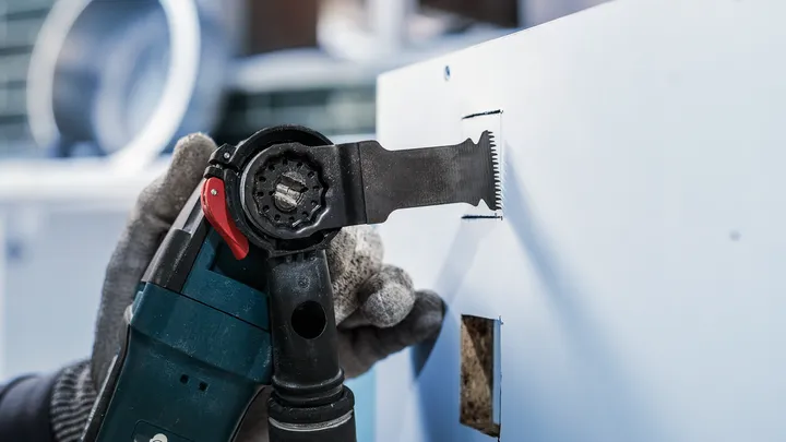 Người mặc đồ bảo hộ đang cắt một lỗ hình chữ nhật trên tường thạch cao bằng dụng cụ điện.