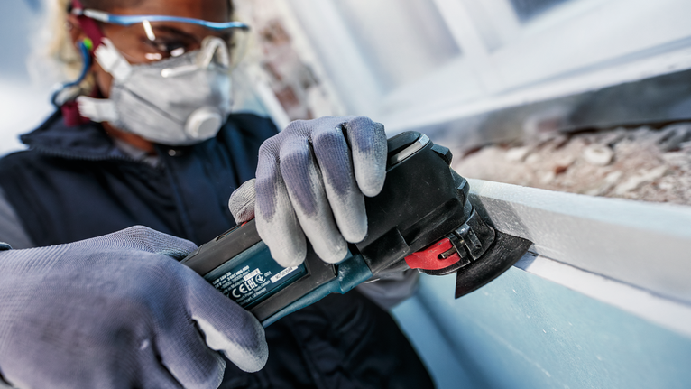 Một người mặc đồ bảo hộ đang mài khung cửa sổ bằng dụng cụ điện.