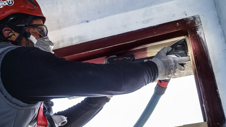 Người đeo thiết bị an toàn đang chà nhám khung cửa sổ bằng gỗ bằng dụng cụ điện.