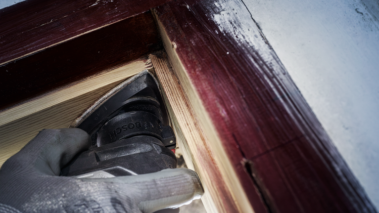 Người mặc đồ bảo hộ đang chà nhám gỗ gần khung cửa sổ đã sơn.