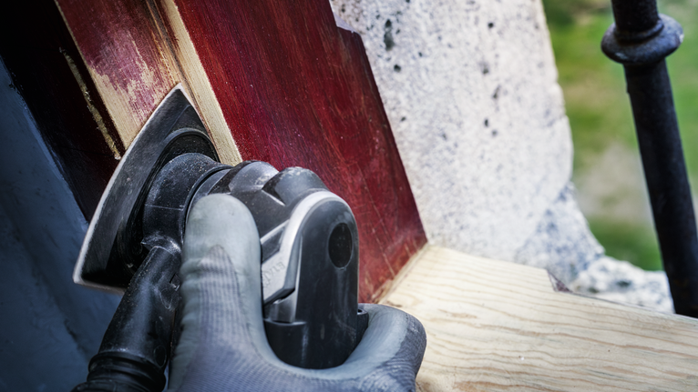 Người đeo thiết bị an toàn đang chà nhám gỗ đã sơn bằng dụng cụ điện.