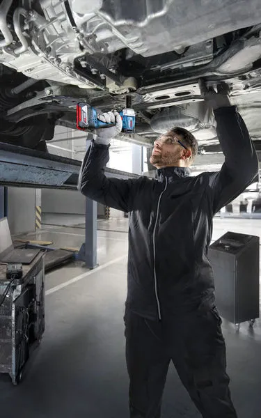 GDS 18V-350 Một người mặc thiết bị an toàn sử dụng máy khoan điện để làm việc bên dưới một chiếc xe đang nâng lên.