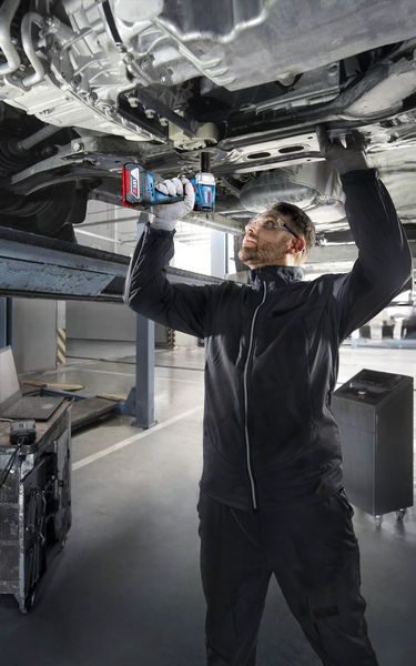 Một người mặc thiết bị an toàn sử dụng máy khoan điện để làm việc bên dưới một chiếc xe đang nâng lên.