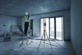 Một người mặc đồ bảo hộ đang điều chỉnh trần nhà bằng dụng cụ cân bằng laser gắn trên giá ba chân.