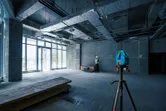Một thiết bị cân bằng laser đặt trên giá ba chân chiếu các đường màu xanh lá cây trong khi một người mặc đồ bảo hộ đang đánh dấu trên tường.