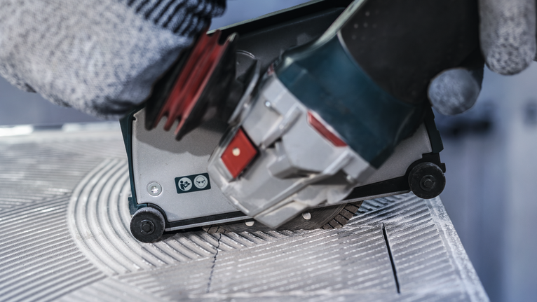 Người đeo thiết bị an toàn đang điều khiển dụng cụ điện cắt rãnh trên gạch.