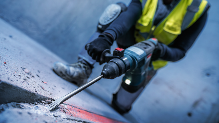 Một người mặc đồ bảo hộ đang đục bề mặt bê tông bằng búa quay.