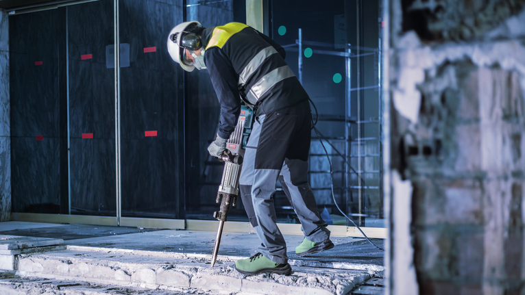 Người mặc đồ bảo hộ đang dùng búa phá bê tông để phá bê tông.