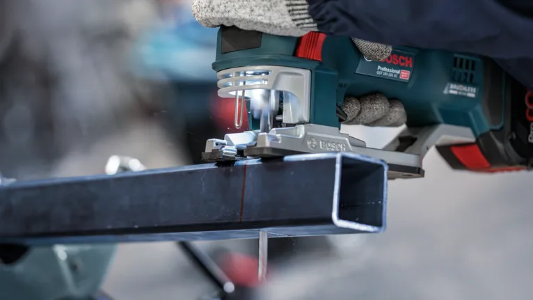 Người mặc đồ bảo hộ đang cắt một thanh kim loại bằng máy cưa điện cầm tay.