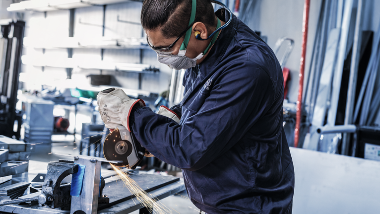 Đá cắt liên kết PRO Stainless Steel and Metal, 125 x 1,6 x 22,23 mm, X-Lock, thẳng, oSa Một người mặc đồ bảo hộ đang mài kim loại, tạo ra tia lửa trong xưởng.