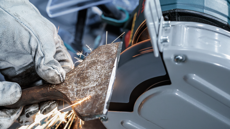 Người đeo thiết bị an toàn đang mài một dụng cụ bằng kim loại trên máy mài.