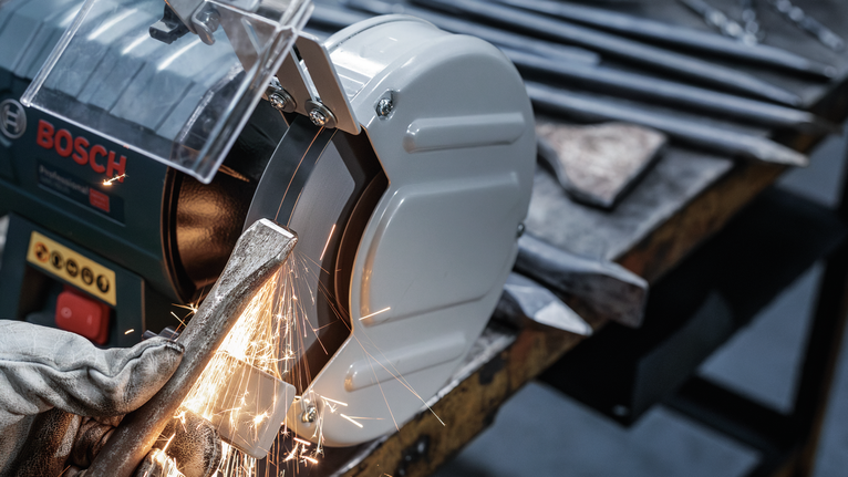 Người đeo thiết bị an toàn đang mài dụng cụ kim loại trên máy mài.