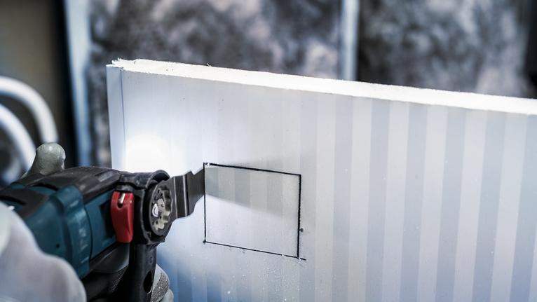 Người đeo thiết bị an toàn đang cắt một lỗ vuông trên vách thạch cao bằng dụng cụ điện.