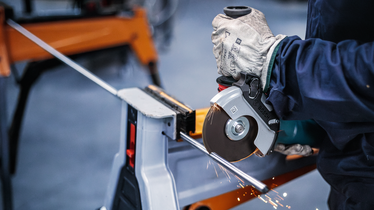 Người đeo thiết bị an toàn đang cắt thanh kim loại bằng dụng cụ điện.