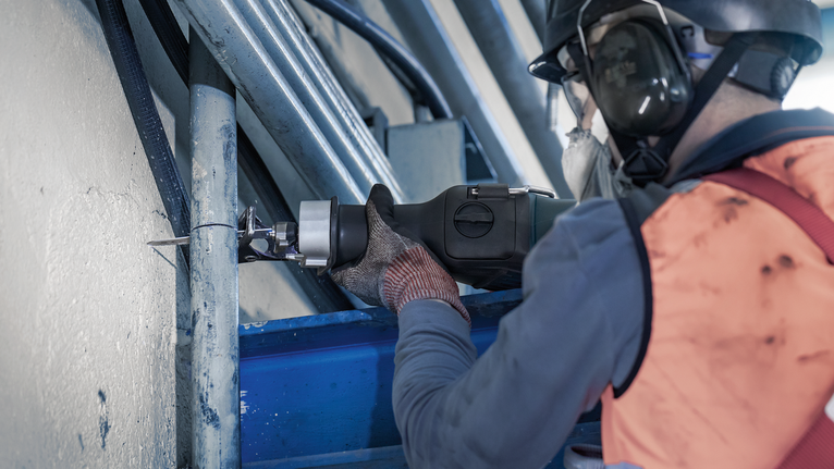 Người mặc đồ bảo hộ đang cắt ống kim loại bằng cưa lọng.