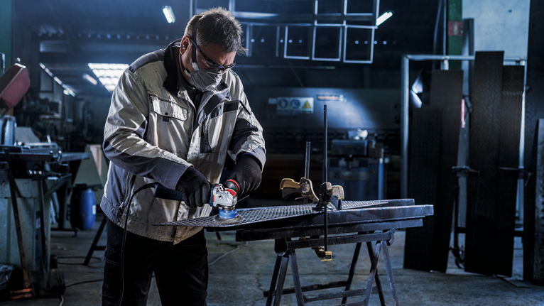 Người mặc đồ bảo hộ đang mài kim loại trên bàn làm việc trong xưởng.