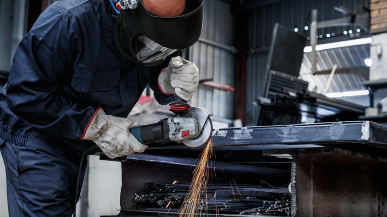 Đĩa mài PRO Metal, 150 x 6 x 22,23 mm Một người mặc đồ bảo hộ đang mài kim loại, tạo ra tia lửa trong xưởng.