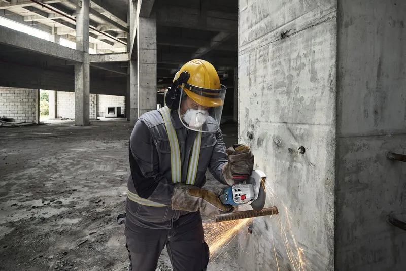 GWS 18V-15 S Một người mặc đồ bảo hộ đang mài thanh thép trên tường bê tông bằng máy mài góc không dây.