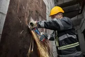 Một người mặc đồ bảo hộ đang mài một tấm kim loại bị gỉ bằng máy mài góc không dây.