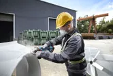 Một người mặc đồ bảo hộ đang mài kim loại ngoài trời tại một khu công nghiệp.