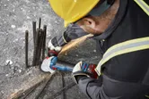 Một người mặc đồ bảo hộ đang mài cốt thép trên bê tông bằng máy mài góc không dây.