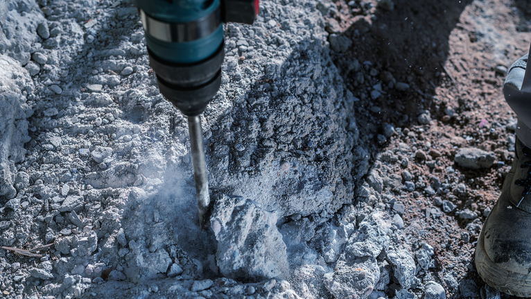 Máy khoan búa đang phá vỡ đống đổ nát bê tông gần một người đang mặc thiết bị an toàn.