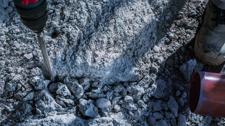 Người mặc đồ bảo hộ sử dụng máy khoan phá dỡ trên nền bê tông thô.