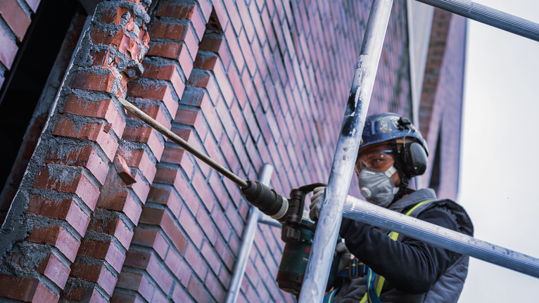 Một người mặc đồ bảo hộ đang khoan vào bức tường gạch trên giàn giáo.