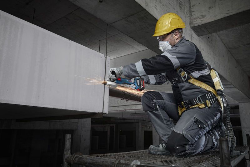 GWS 18V-8 Một người mặc đồ bảo hộ đang mài bê tông bằng dụng cụ điện tại công trường xây dựng.