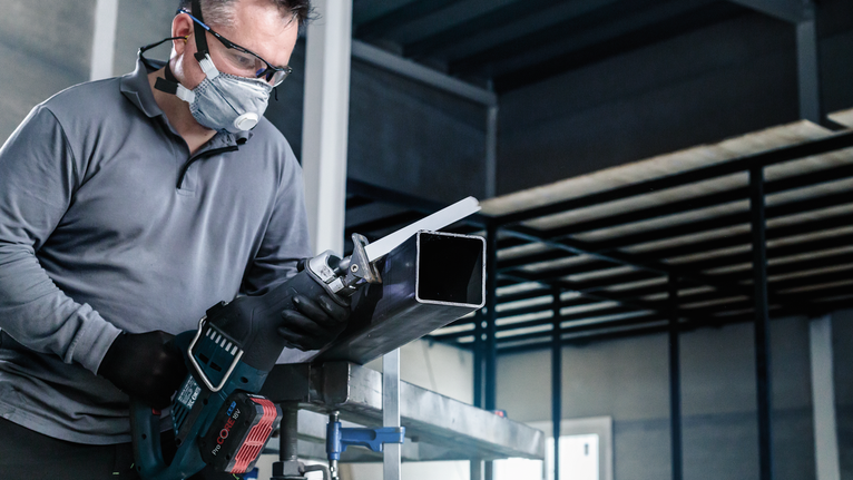 Một người mặc đồ bảo hộ đang cắt một ống kim loại bằng cưa lọng.