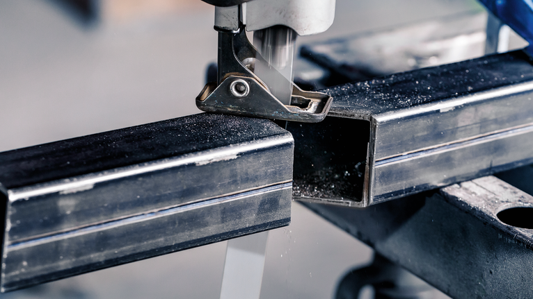 Máy cưa lọng đang cắt một ống kim loại hình chữ nhật trên bàn làm việc.