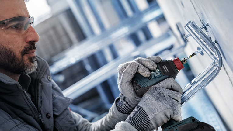 Một người đàn ông mặc đồ bảo hộ đang dùng máy khoan điện để bắt vít giá đỡ vào tường.