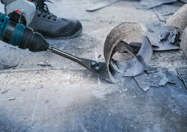 Một người mặc đồ bảo hộ đang sử dụng dụng cụ điện để cạo vật liệu sàn.