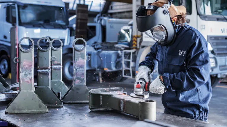 Người mặc đồ bảo hộ đang mài một bộ phận kim loại trong môi trường công nghiệp.