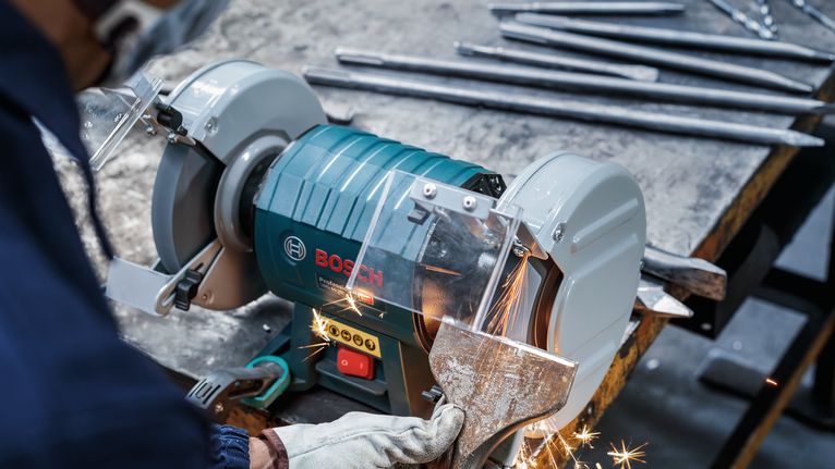 Một công nhân mặc đồ bảo hộ đang mài một miếng kim loại bằng máy mài bàn.