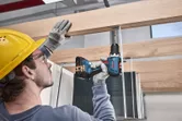 Một người đeo thiết bị an toàn sử dụng máy khoan không dây để vặn vít vào thanh gỗ.