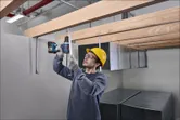 Một người mặc thiết bị an toàn sử dụng máy khoan điện để cố định các giá đỡ bằng kim loại ở phía trên.